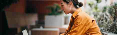 A woman in an orange shirt works on a laptop at an outdoor table.